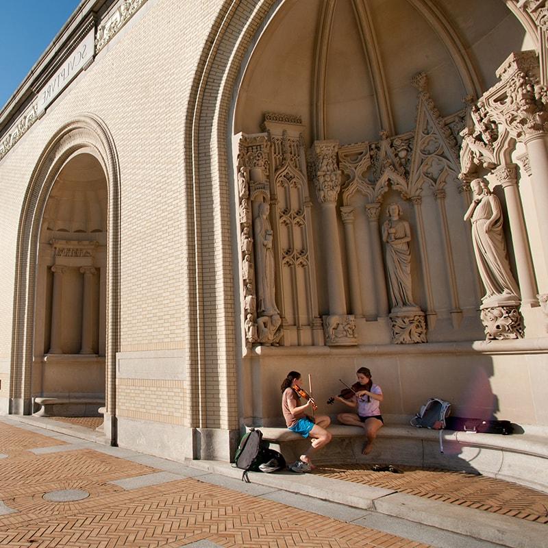 Photo of a campus building with two students playing violin